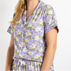 A smiling blonde woman wearing a short-sleeve lavender pajama shirt with a white floral print and white piping, paired with a matching drawstring waist, against a plain white background.