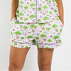 A person wearing a matching short-sleeve pajama top and shorts set with a white background and an all-over colorful cartoon print of trees, birds, and houses, plus purple piping details.
