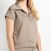 A woman wearing a taupe short-sleeve half-zip pullover top, shown from the waist up against a clean white studio background.