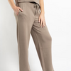 A person wearing loose, straight-leg lounge pants in a warm taupe-beige color, paired with a matching top. The photo is cropped from the chest down, showing the model's hands in the pockets and bare feet against a clean white studio background.