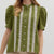 Woman wearing a green blouse with white lace details and beige pants on a white background